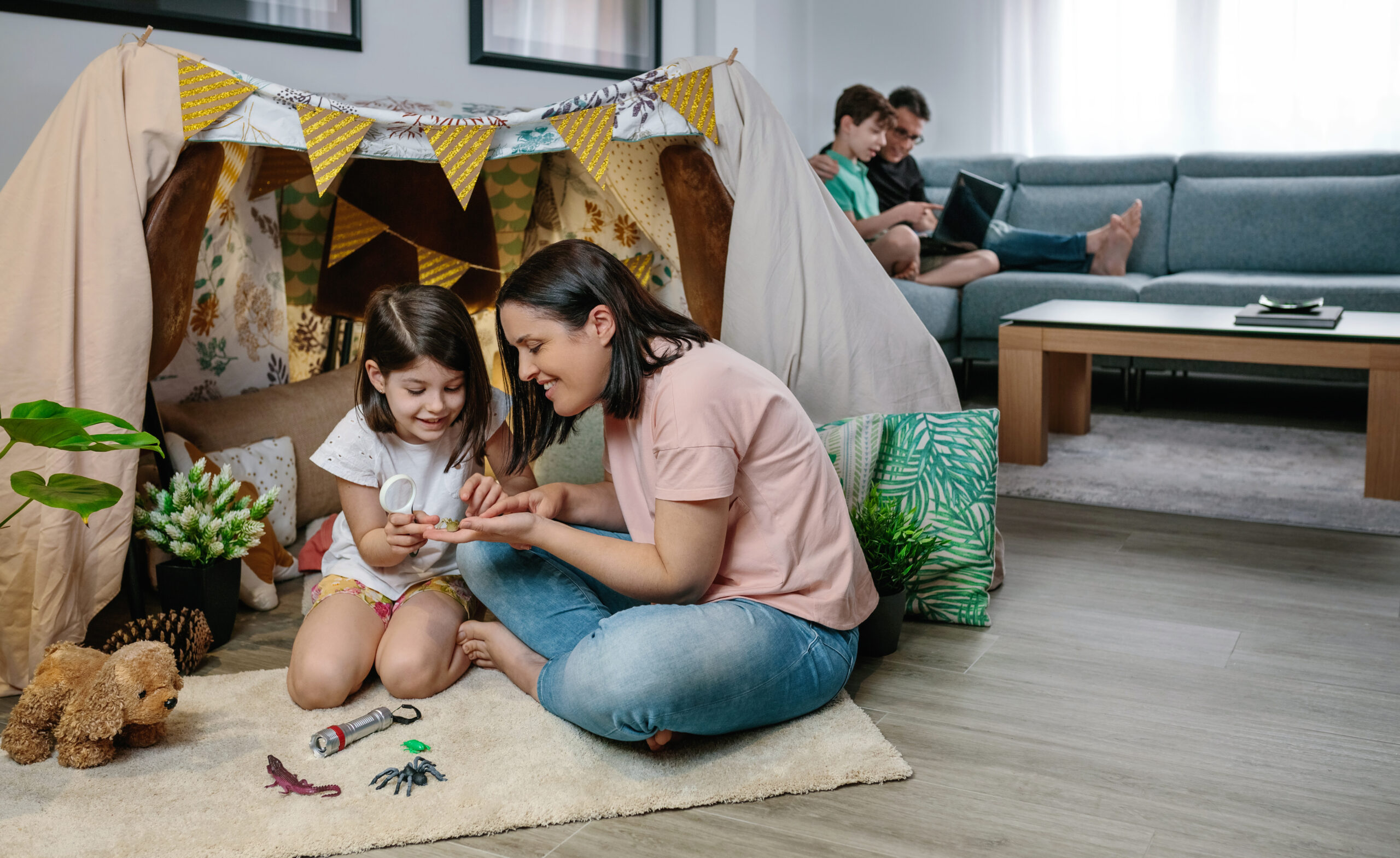Mother and daughter play camping at home while father and son use laptop on the sofa
