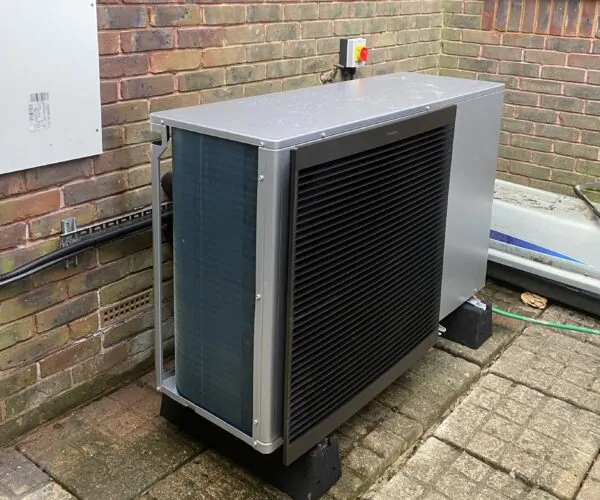 Outdoor heat pump or air conditioning unit mounted on black supports beside a brick wall, with visible grilles, piping, and an electrical isolator switch above it.