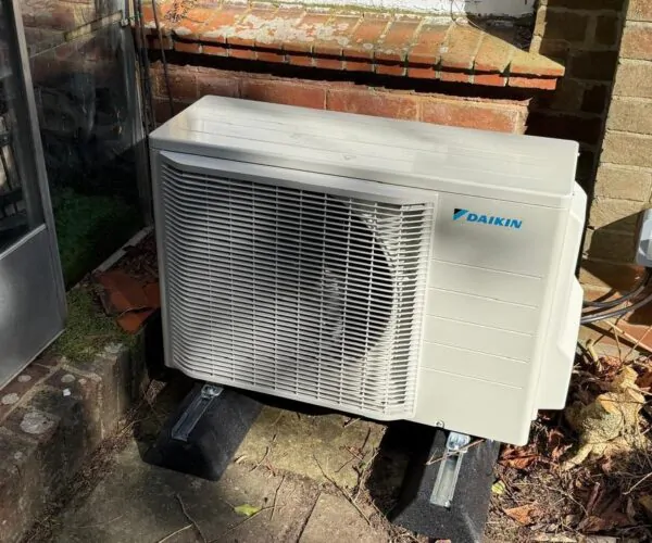 Daikin outdoor air conditioning or heat pump unit mounted on black supports beside a brick wall and patio area.