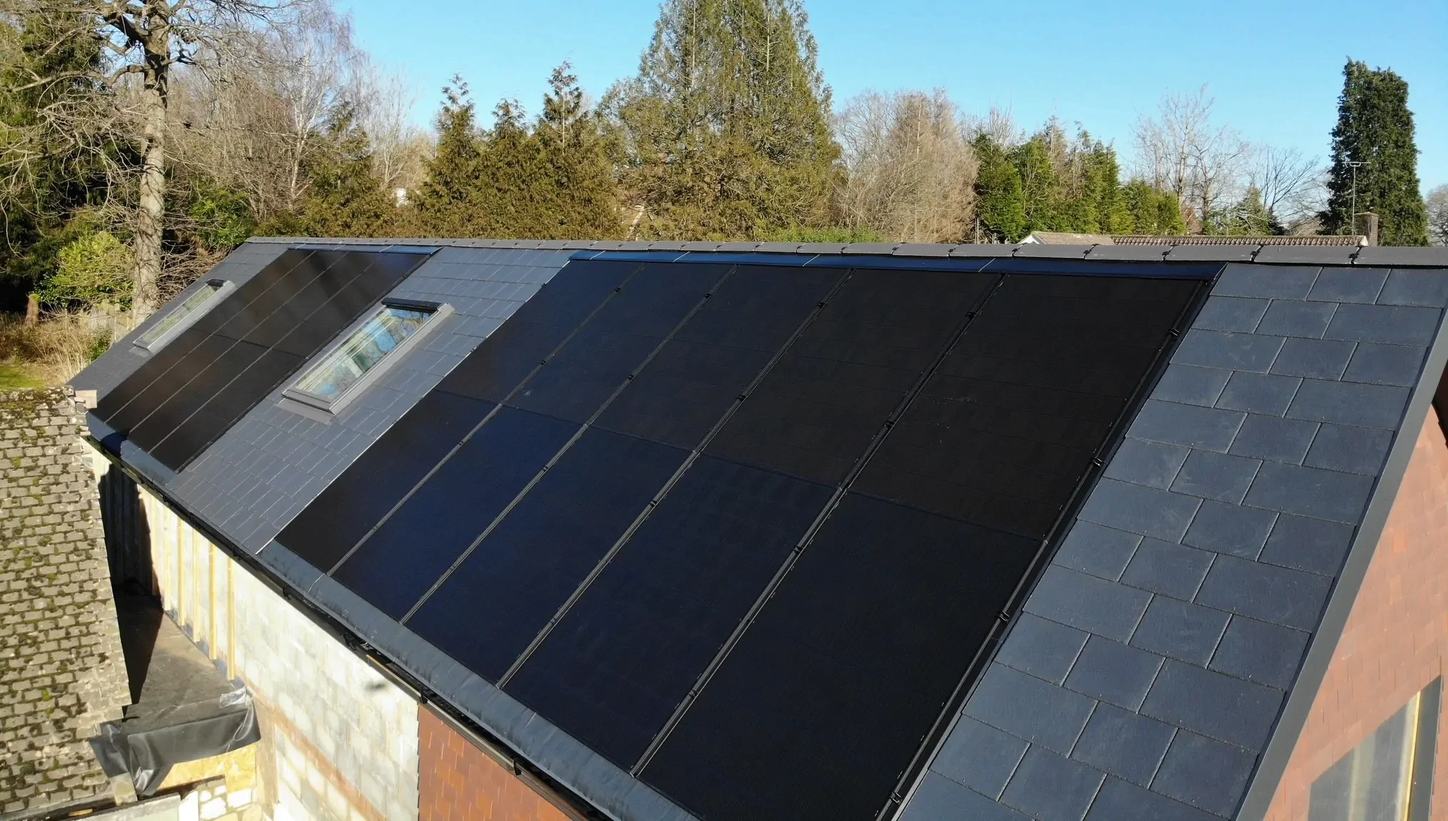 Slate roof fitted with large black solar panels and a skylight, with trees and blue sky in the backgroun